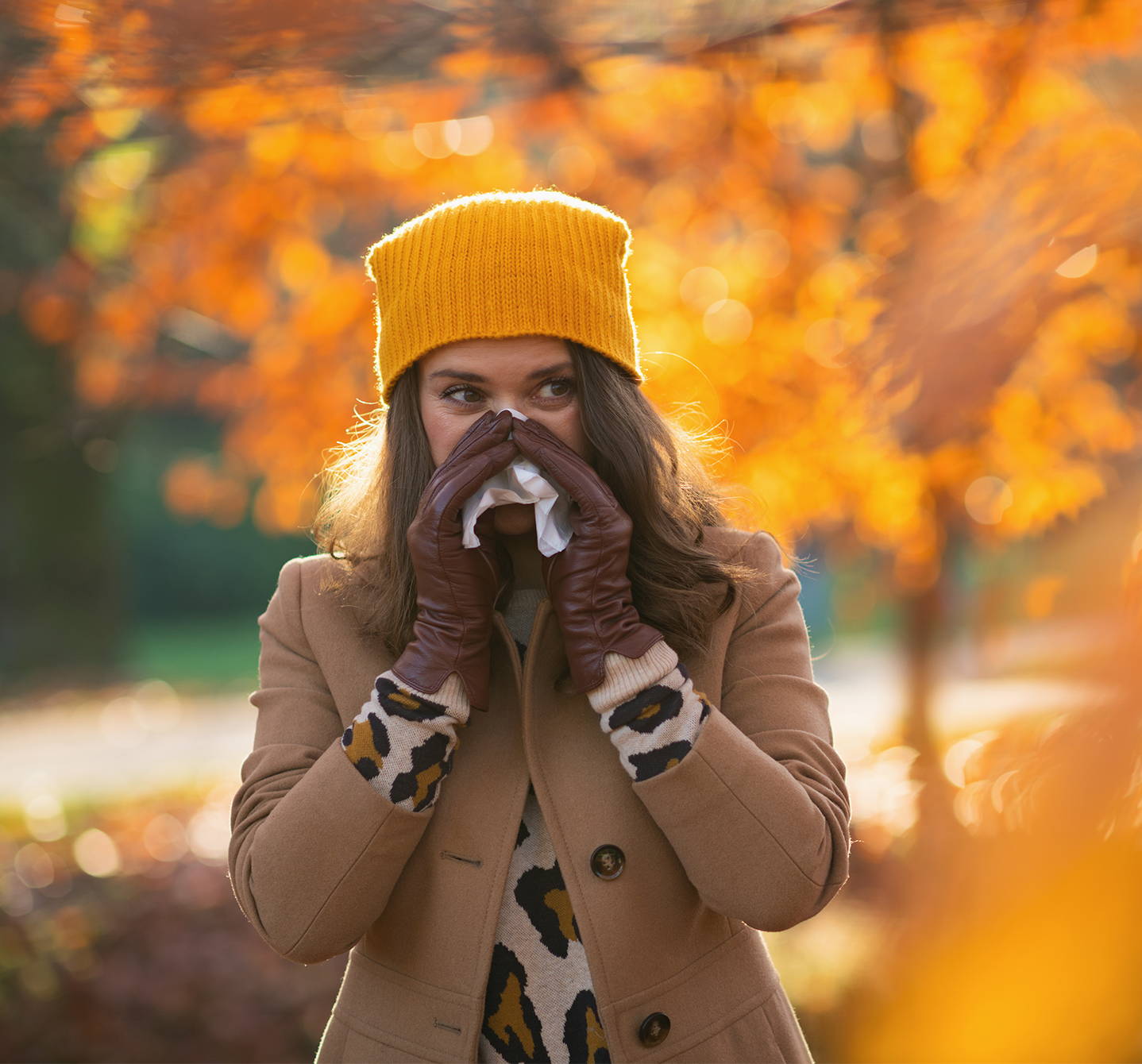  Symptome einer Schimmelpilzallergie – Frau mit Kopfbedeckung und Handschuhen, die sich die Nase putzt als Reaktion auf Schimmel, der sich in Herbstlaub bildet.