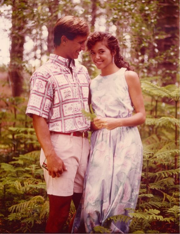 A couple standing together in a forest, smiling at each other.