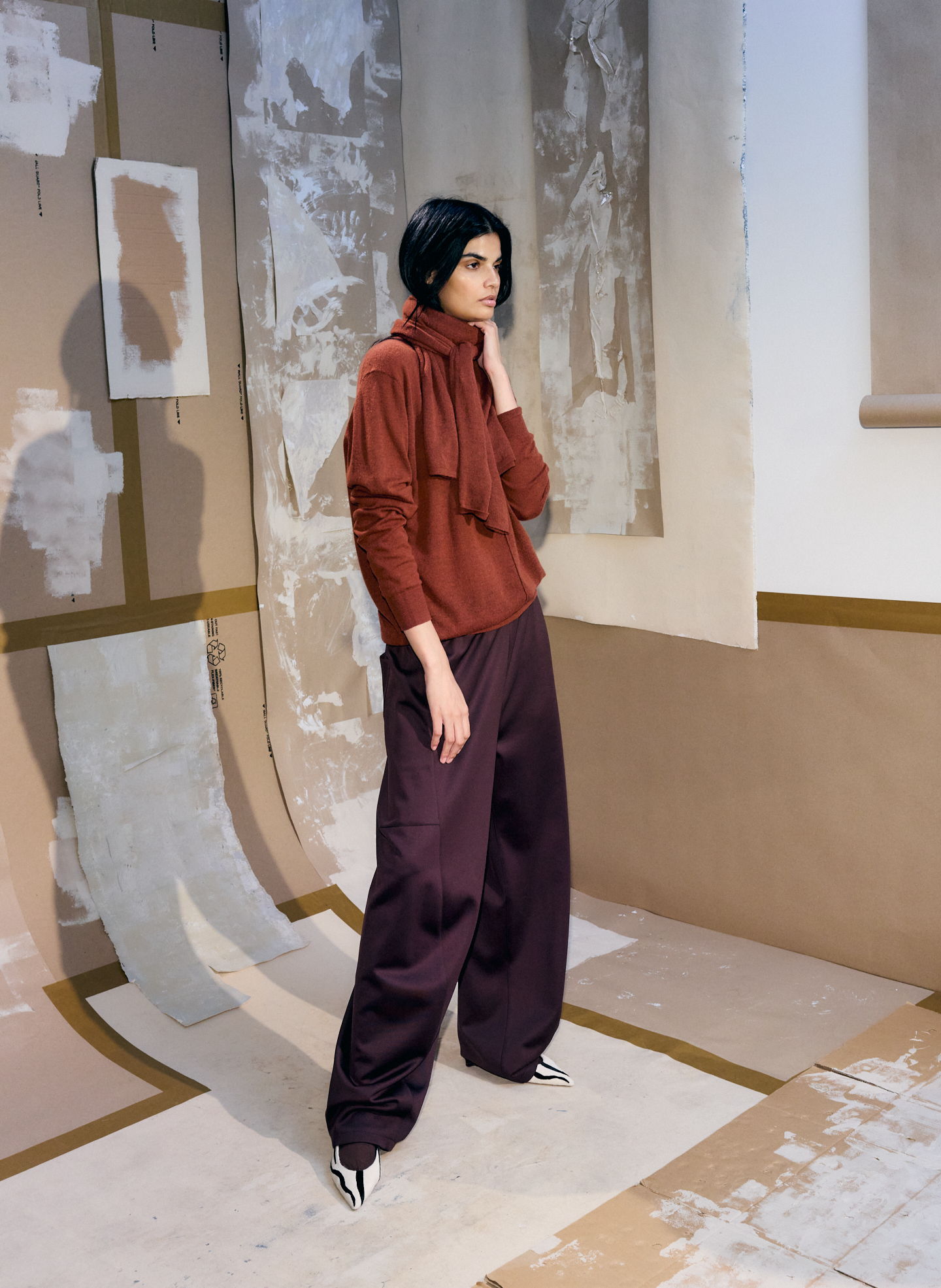 Model wearing a rust-colored top and purple pants in an artistic setting.