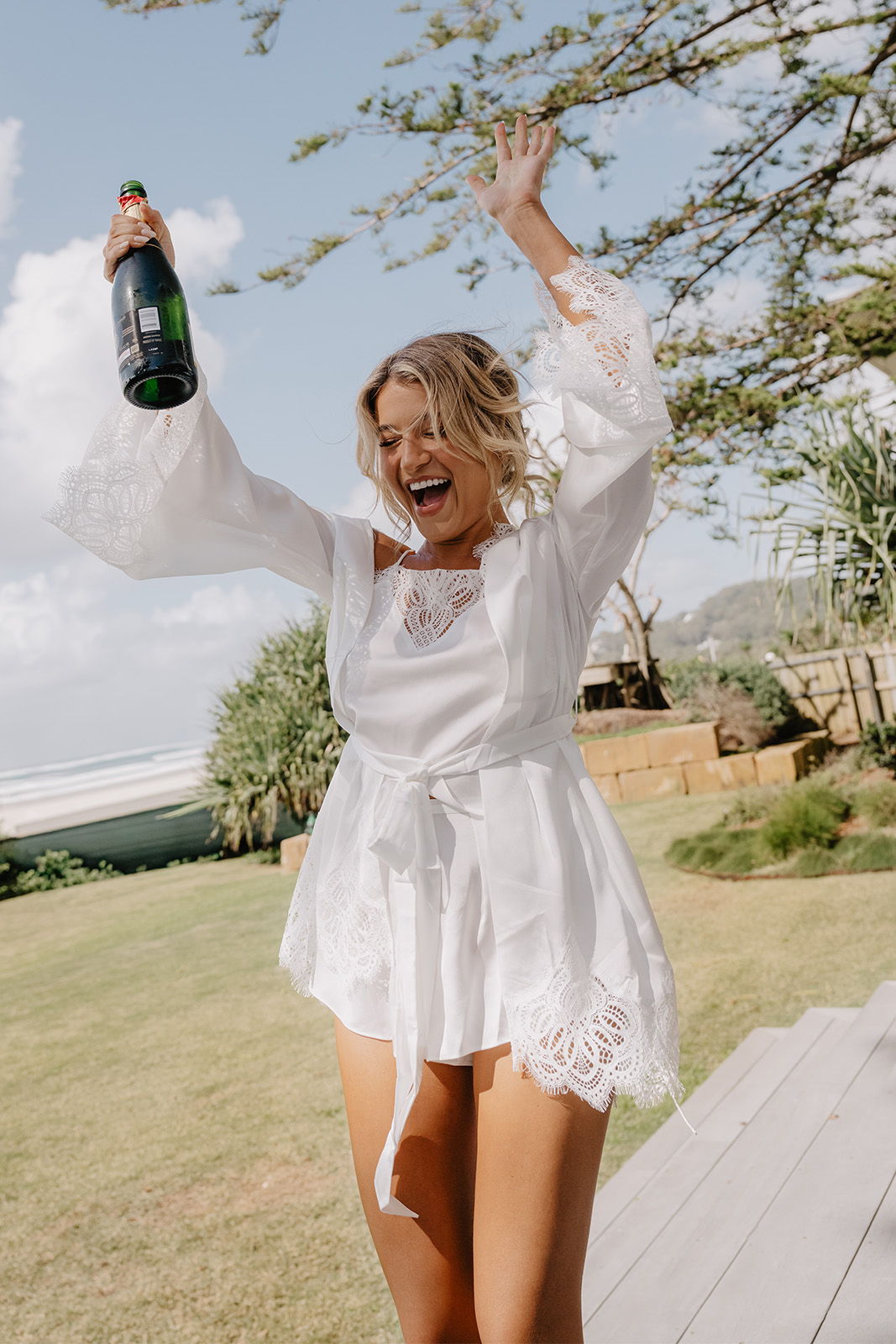Woman in a white dress celebrating with champagne outdoors.