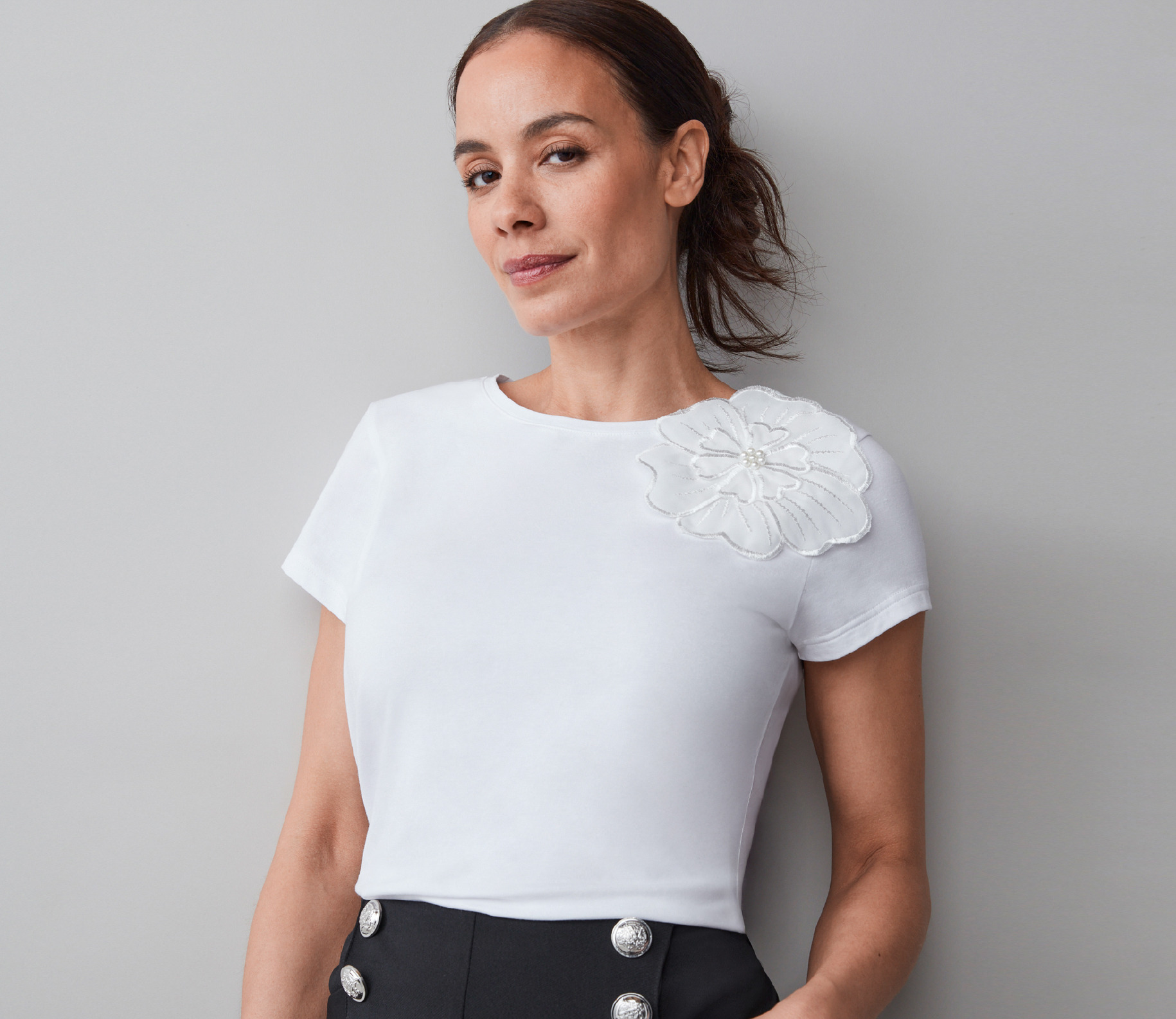 Woman in a white top with a floral detail, standing against a gray background.