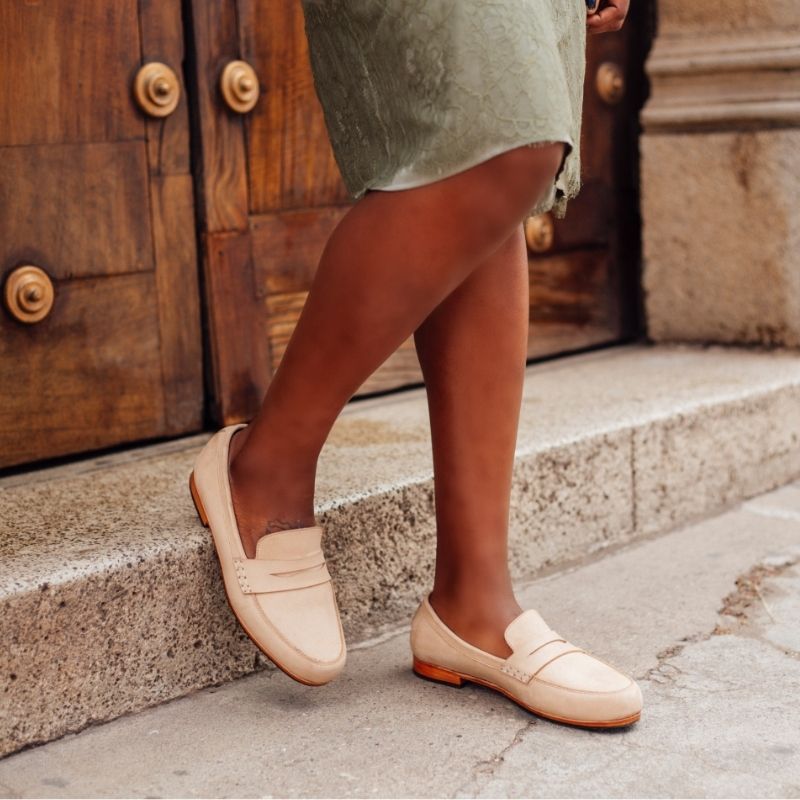 Side lifestyle view of a woman wearing the handcrafted Adelante Sabina women's wide width leather loafers in a soft cream oatmeal tone, stepping down a stone curb near a large wooden door.
