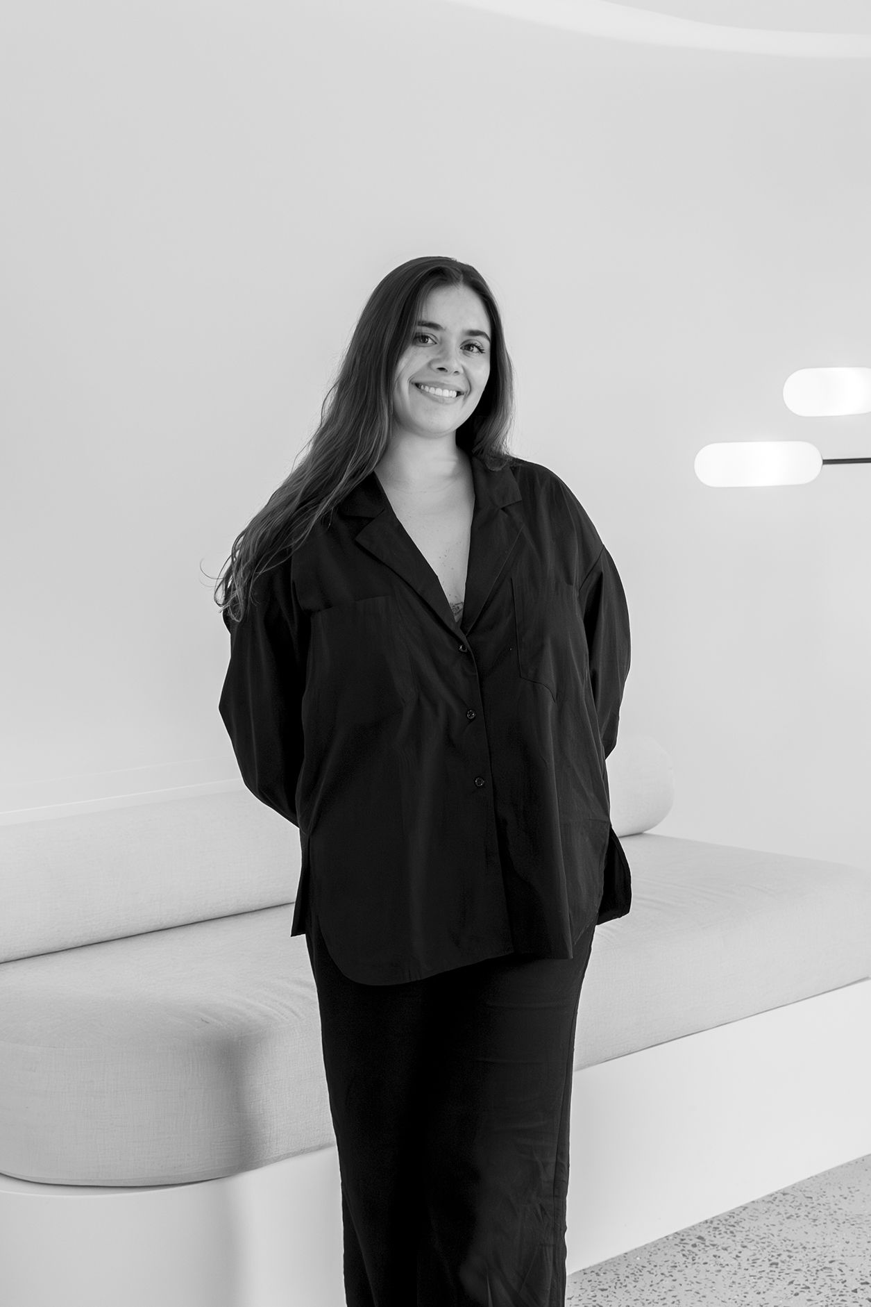 A woman in a black outfit stands smiling in a modern interior.
