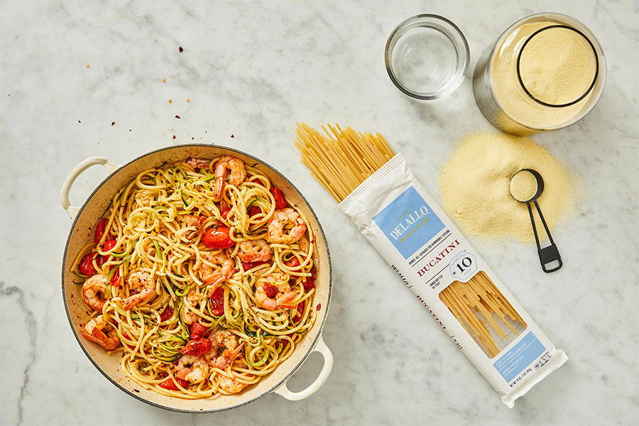 A prepared pot of bucatini pasta tossed with tomatoes and shrimp set next to a package of DeLallo Bucatini Pasta