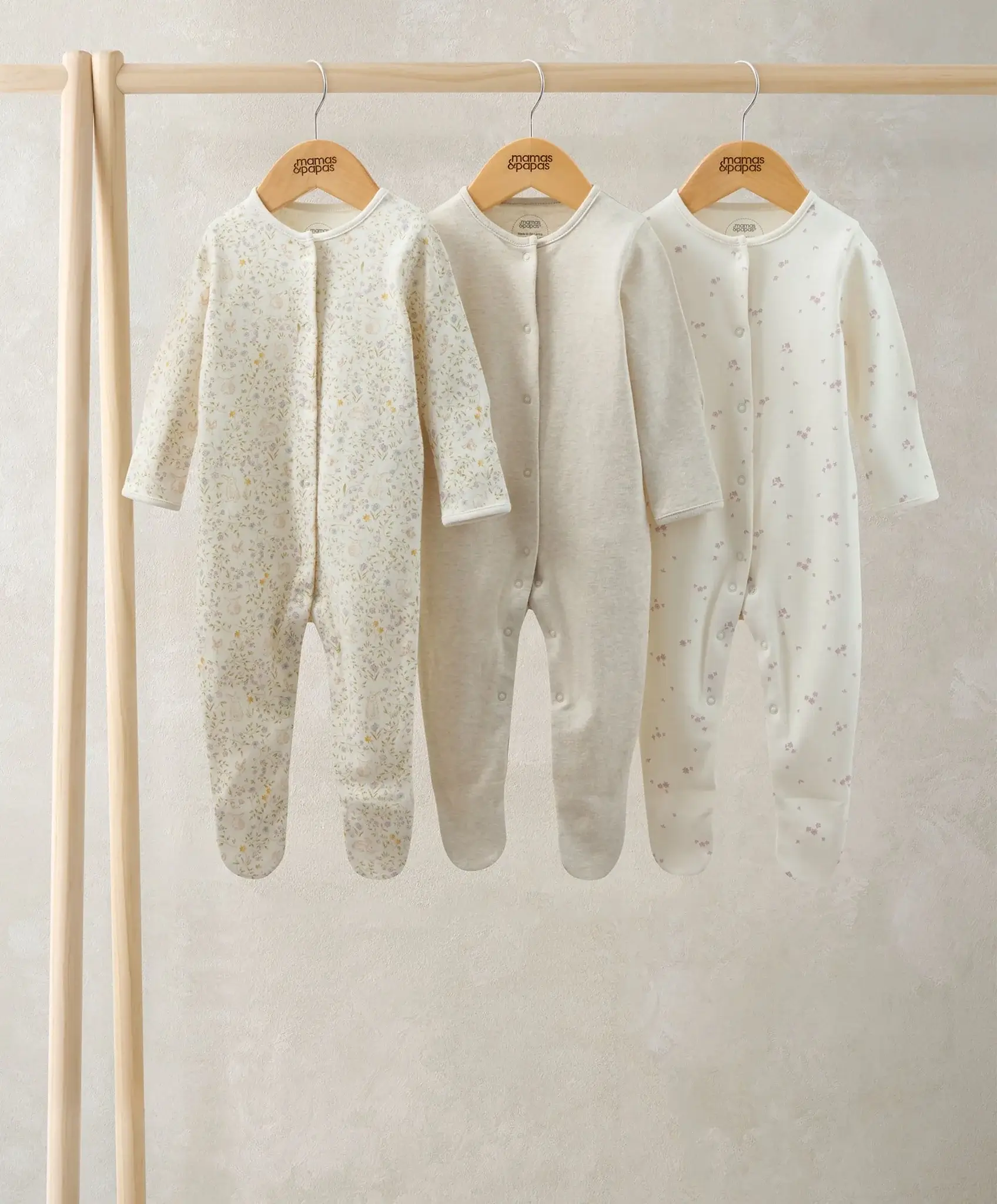 Three baby onesies hanging on wooden hangers against a neutral background.