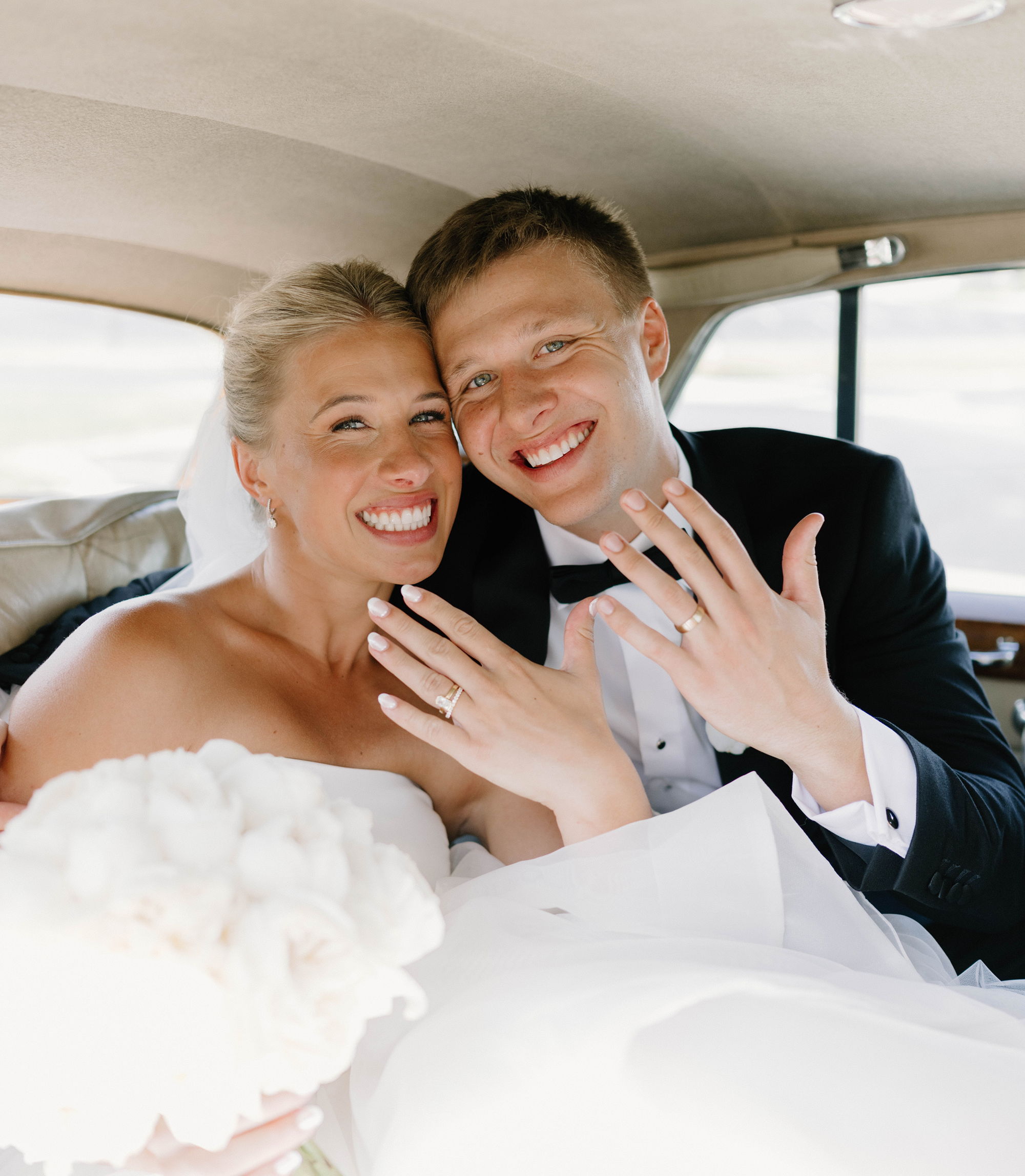 Bride and groom showing wedding rings inside a car, smiling happily.