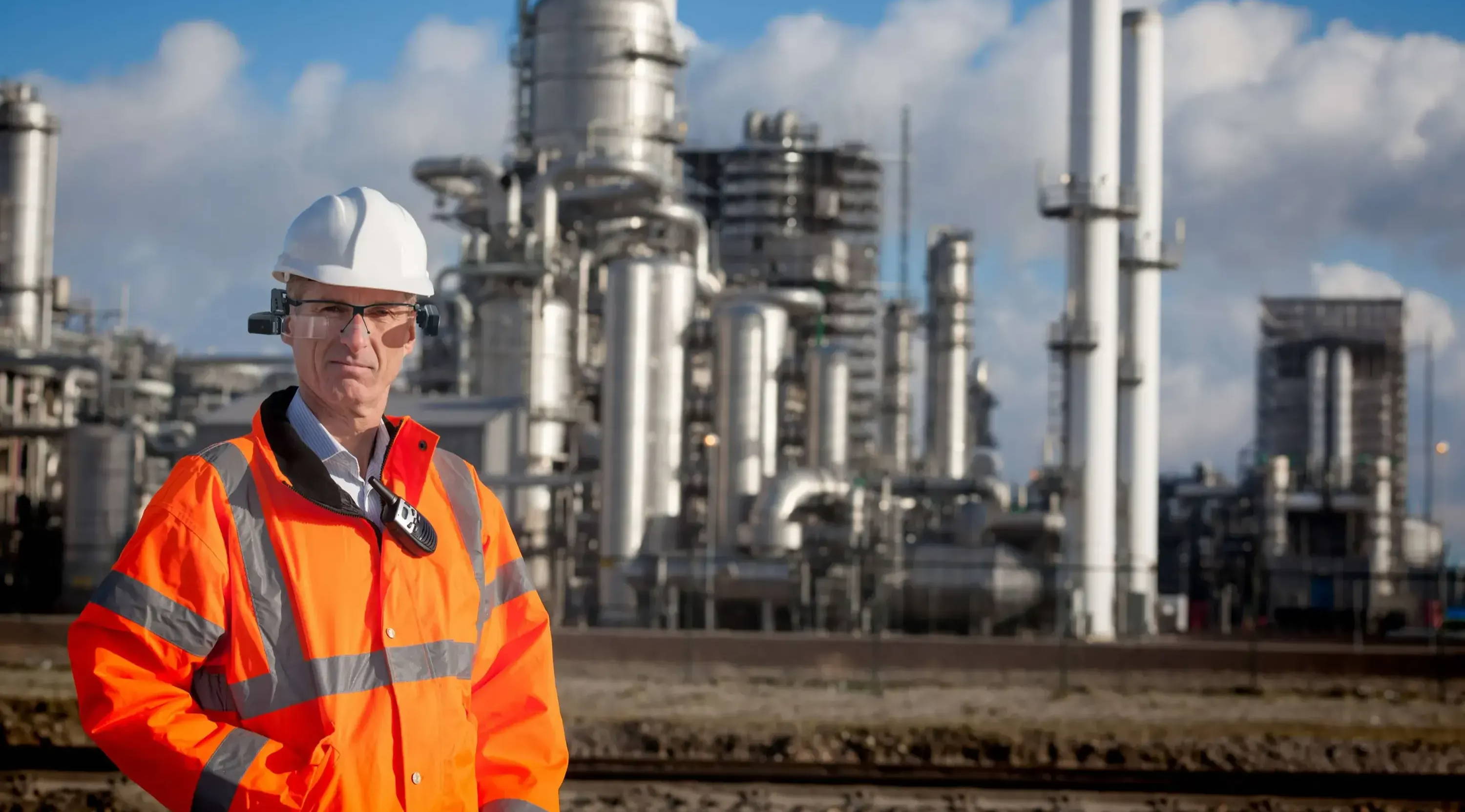 Man standing in front of a processing plant, wearing Vuzix smart glasses, white hard hat, and orange safety jacket. 