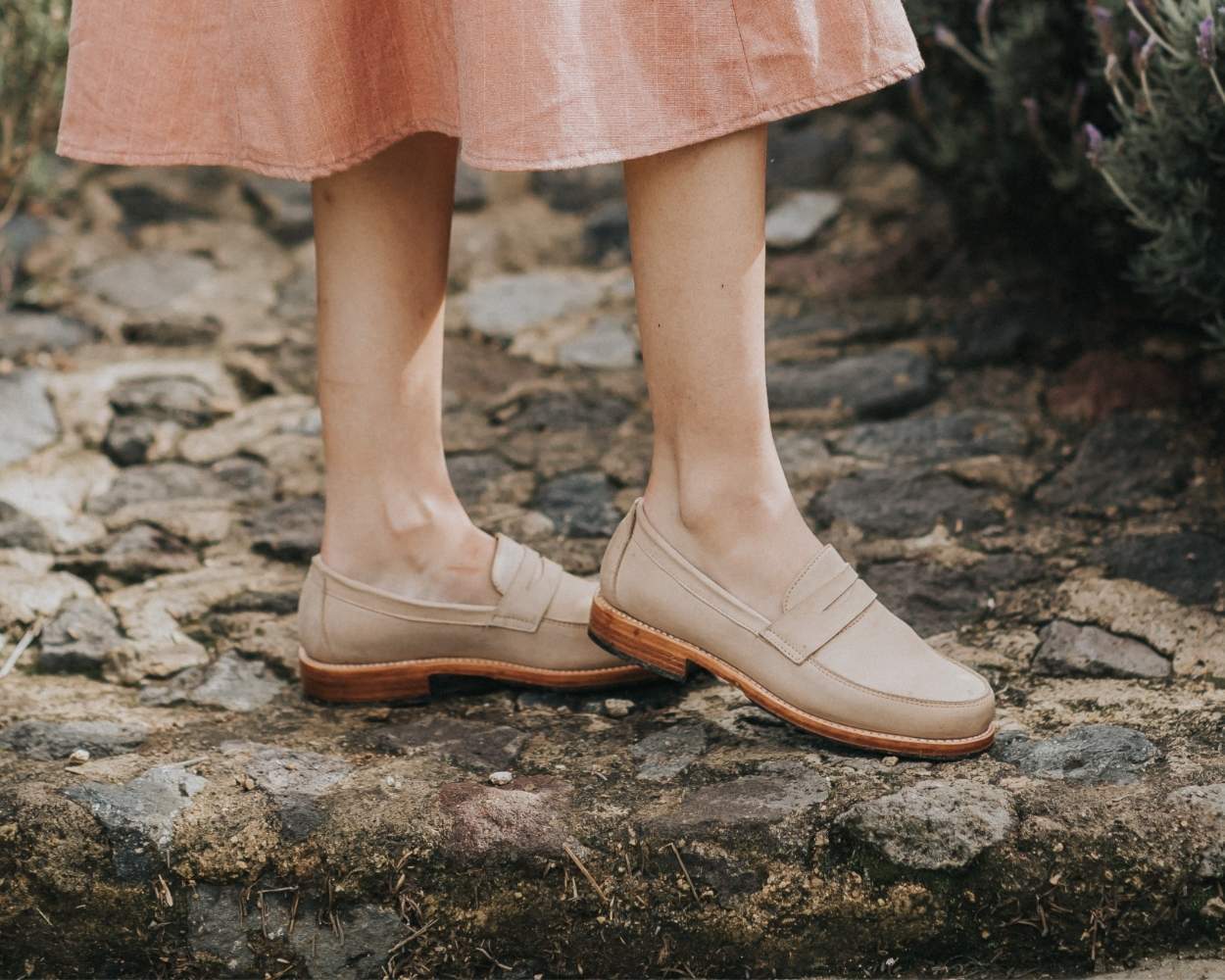 An outdoor lifestyle shot from the calves down of a person standing on a rustic, uneven stone path. They are wearing a light pink or peach midi skirt and a pair of classic beige or cream leather penny loafers featuring distinct visible stitching and a low, stacked light brown leather sole.