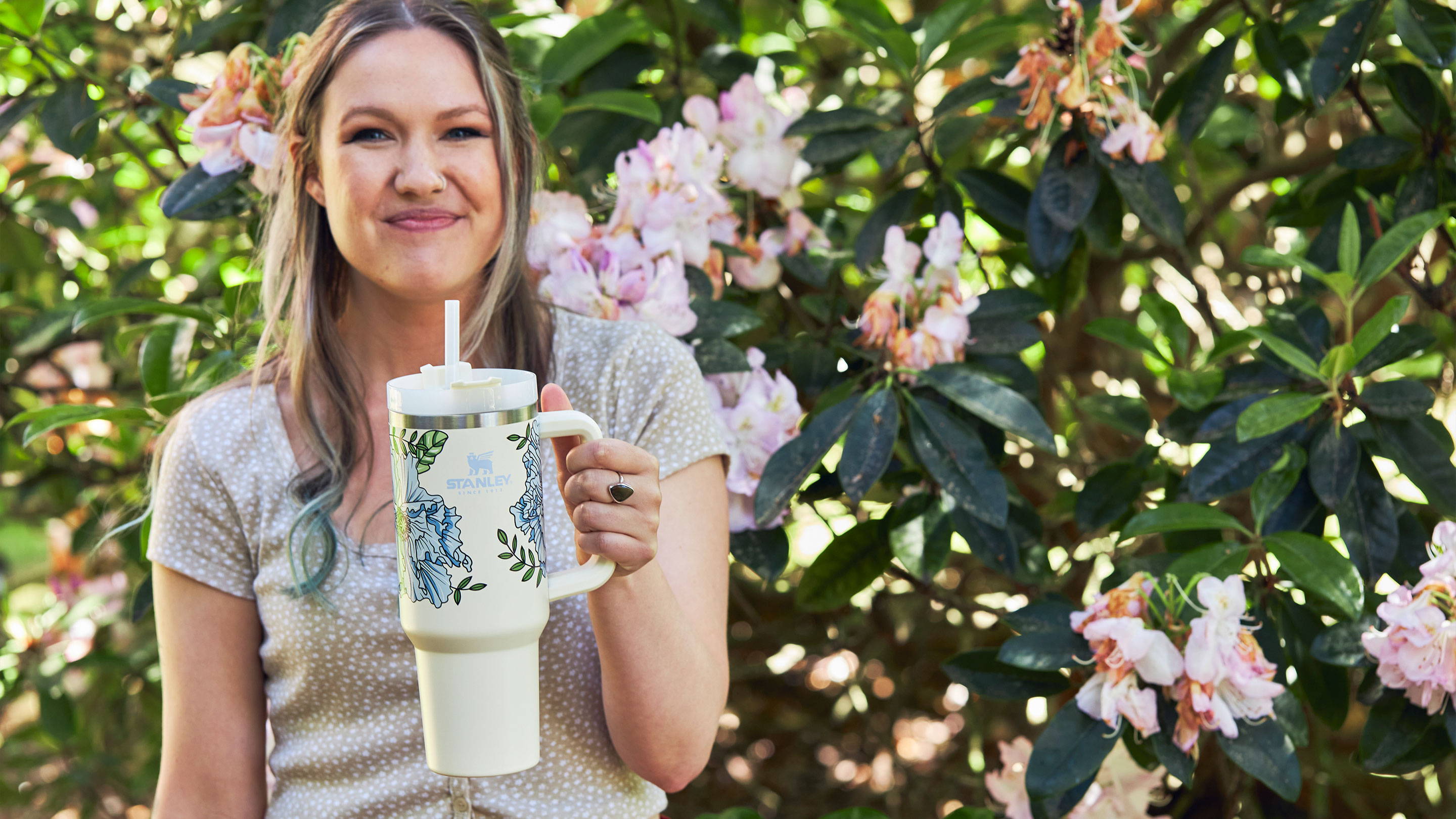 Glynn Rosenberg stands in front of a rhododendron with pink flowers holding the 40-oz insulated Adventure Quencher Travel Tumbler in Cream Floral.