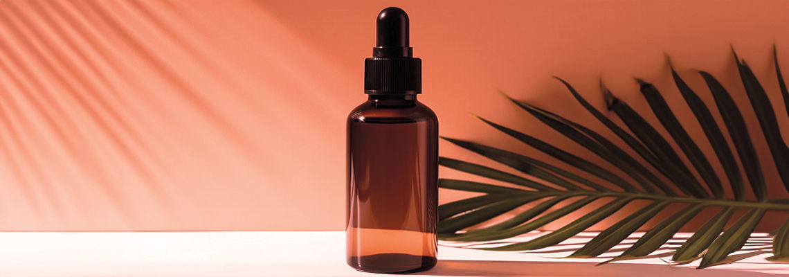 Brown glass dropper bottle against a soft pink background with a palm leaf shadow.