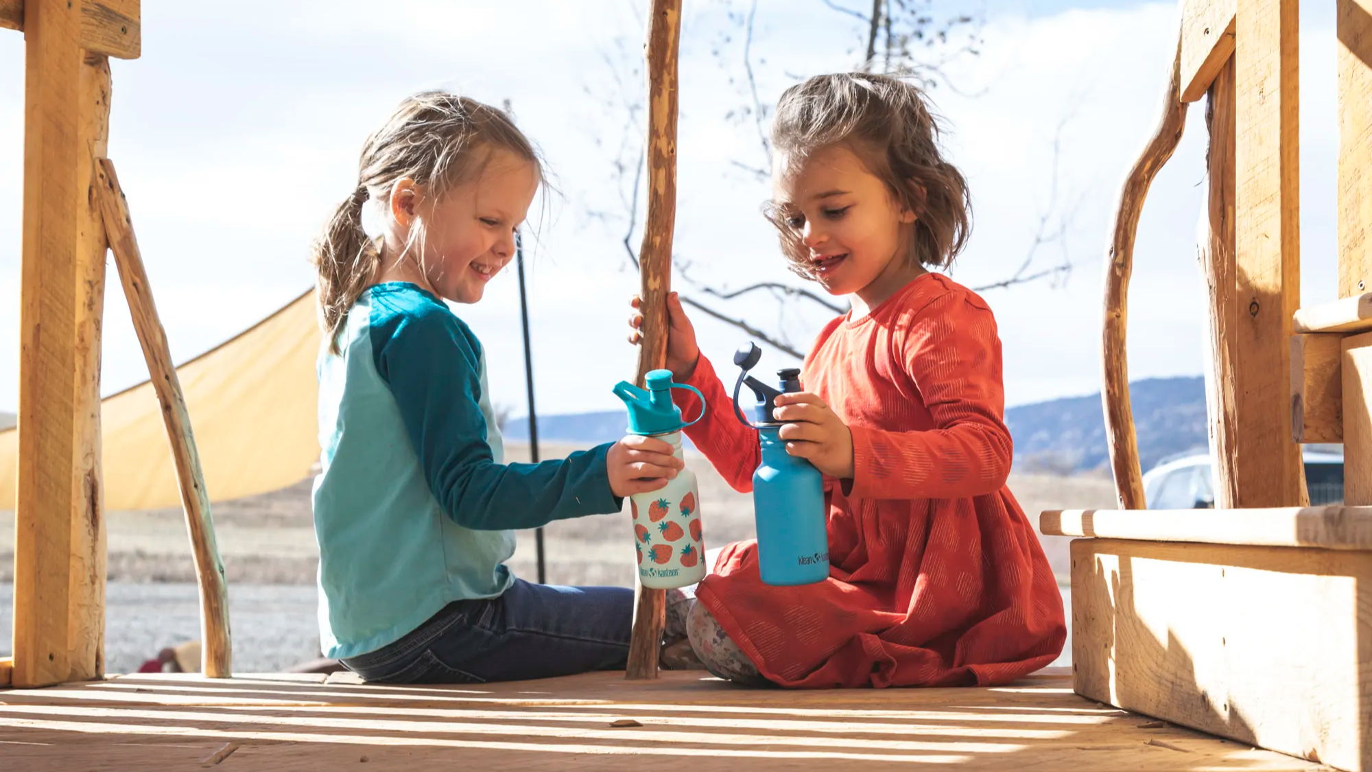 two kids sitting on a wooden play structure with two kid kanteen bottles with a sports cap