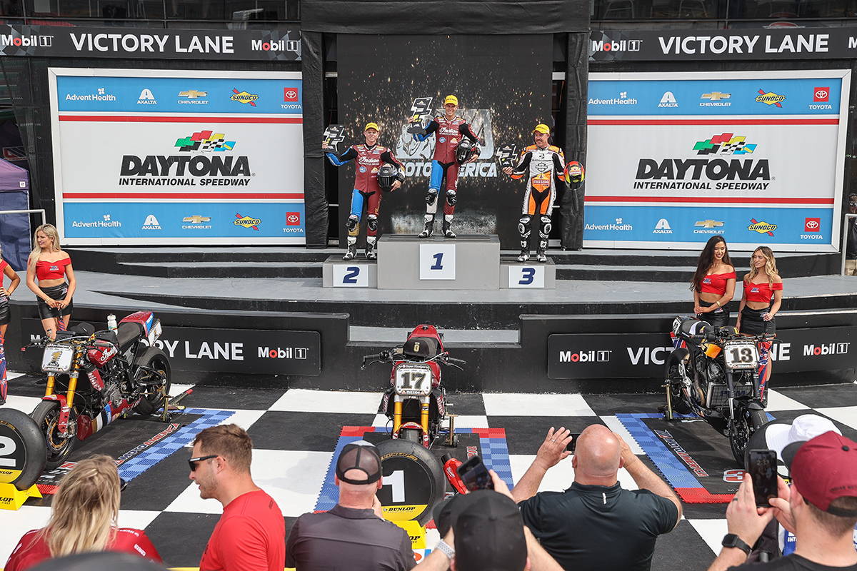 Race winner in a maroon Indian Motorcycle racing suit celebrates on the podium, surrounded by team members and race umbrellas, giving a peace sign with a joyful expression.