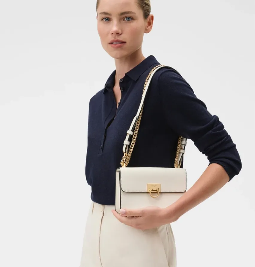 A woman holding a white handbag, wearing a navy shirt and light pants.