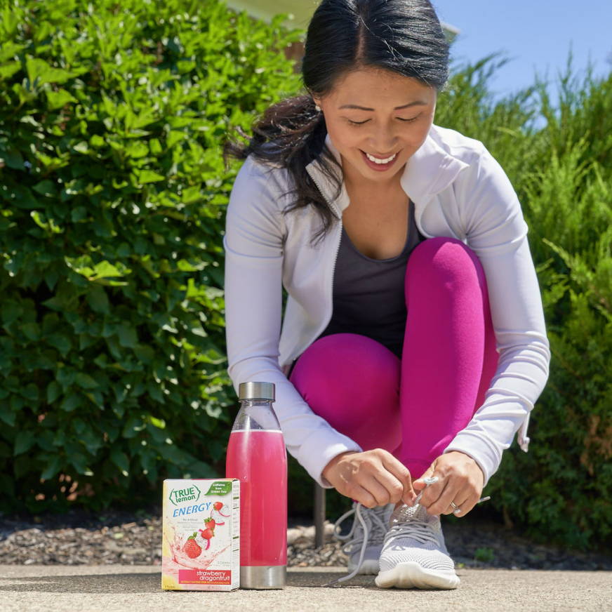 Woman kneeling down to tie her running shoes. Right next to her is a box of true lemon energy strawberry dragonfruit packets and a bottle of the flavored water.