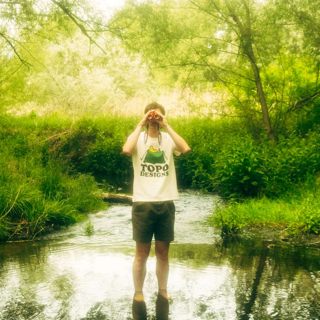 Person standing in a stream, holding a camera, surrounded by greenery.