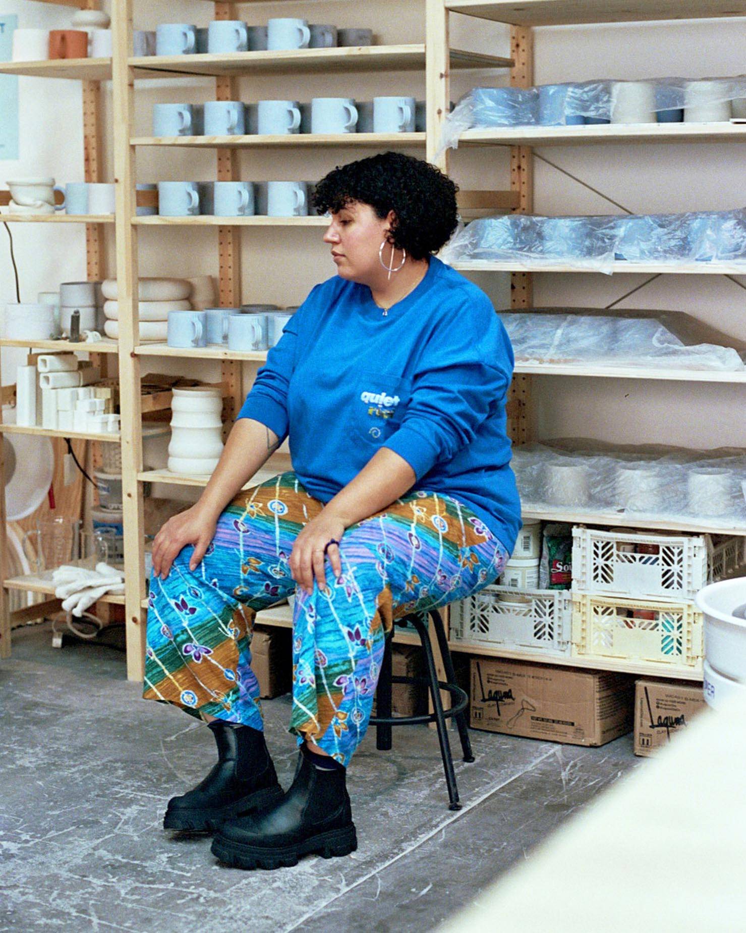 Sara sitting in her ceramic studio wearing a blue shirt by Martine Rose and pants from Collina Strada. 