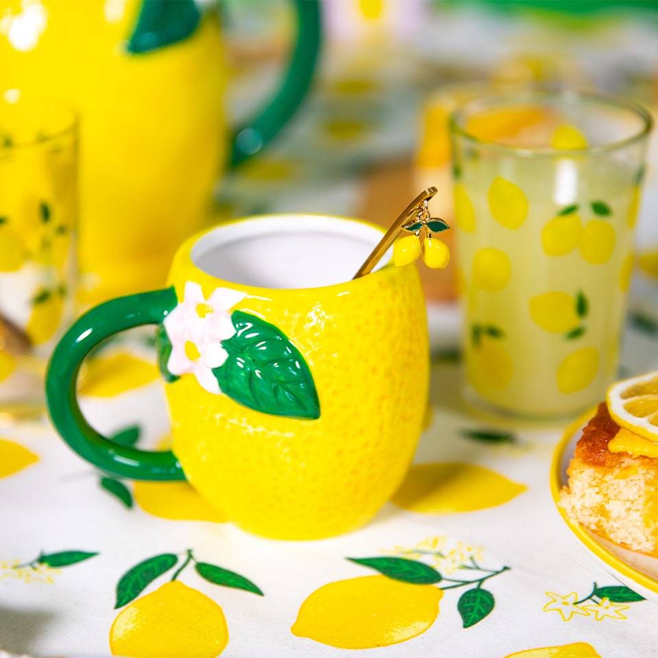A close-up of a vibrant lemon mug with a green handle, decorated with a small flower, placed on a tablecloth with lemon prints next to a glass of lemonade and a slice of lemon cake on a wooden board.