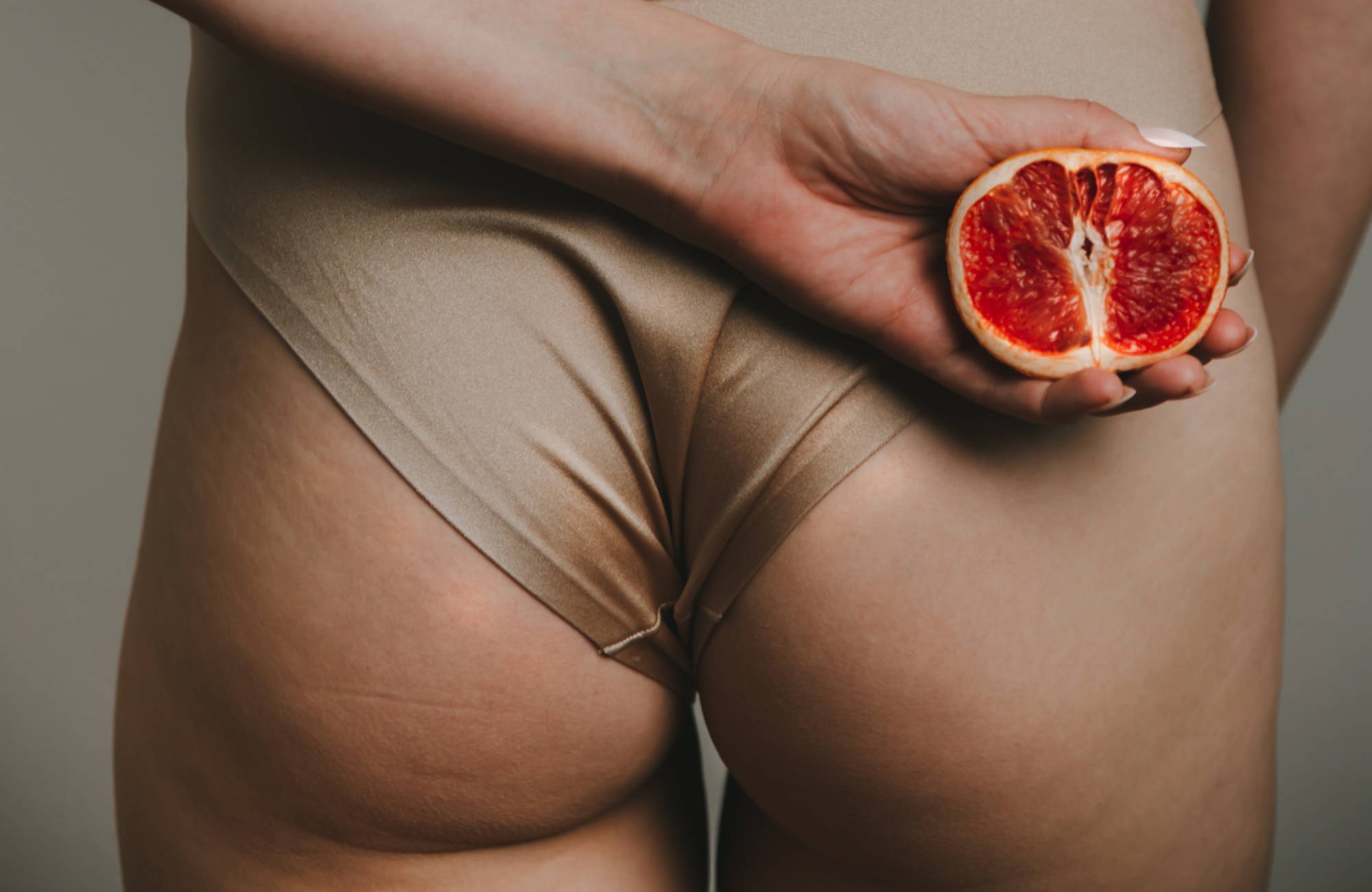 a woman’s butt in a nude bodysuit holding a vulva-esque fruit behind her back