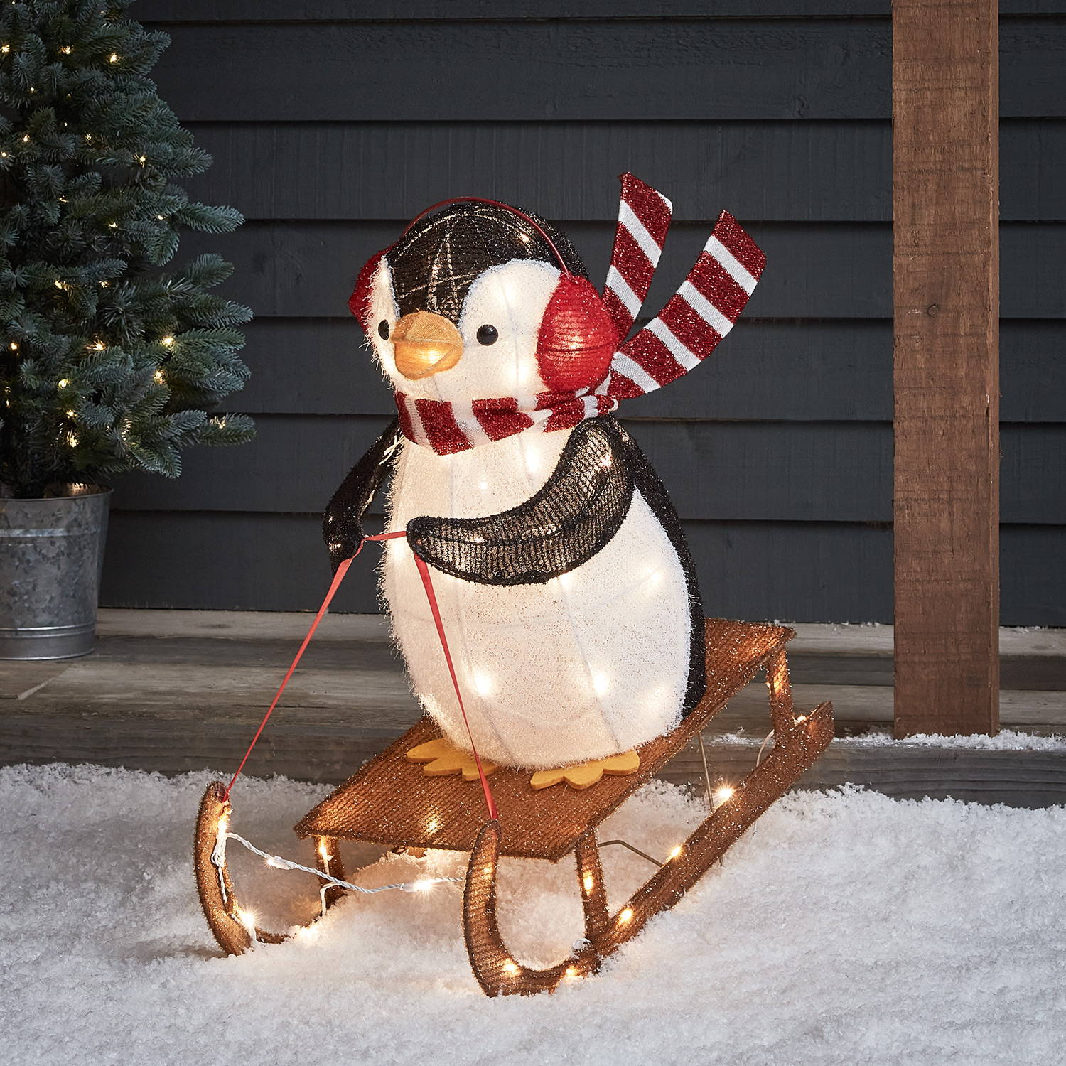 Sledging penguin sliding off a porch onto a snowy garden outside a cosy cottage 