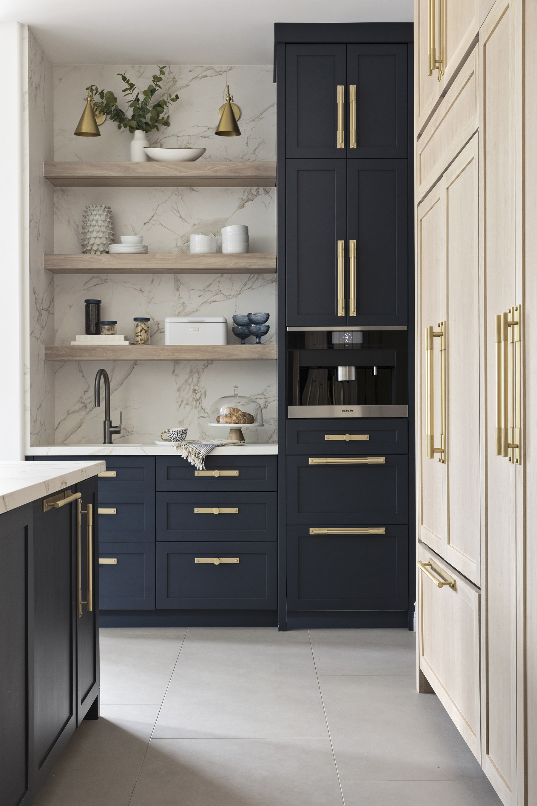 Navy and light wood kitchen complete with brass cabinet hardware, marble worktops, tiled floor and wall shelves