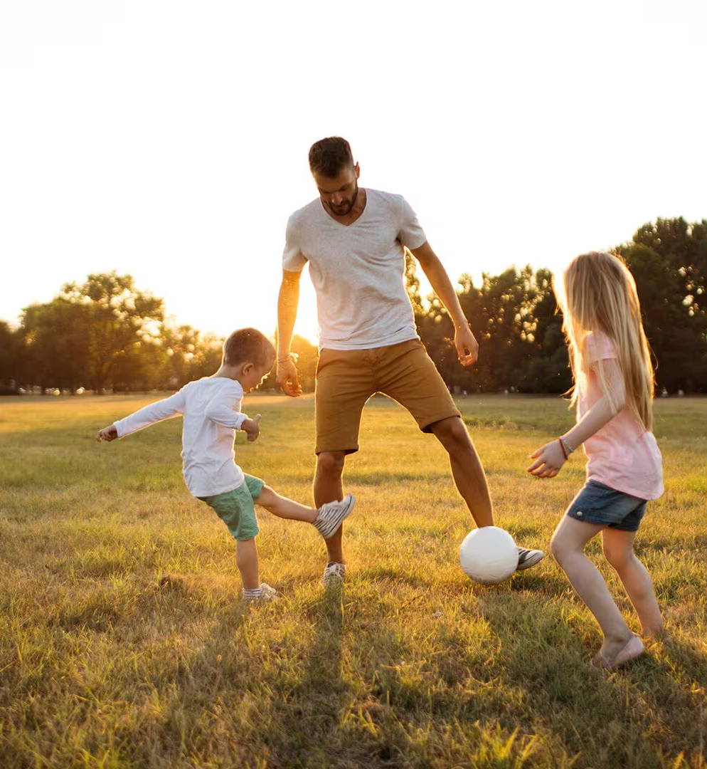 Otec hrajúci futbal na tráve so svojím malým synom a dcérou v zlatistom svetle neskorého popoludnia