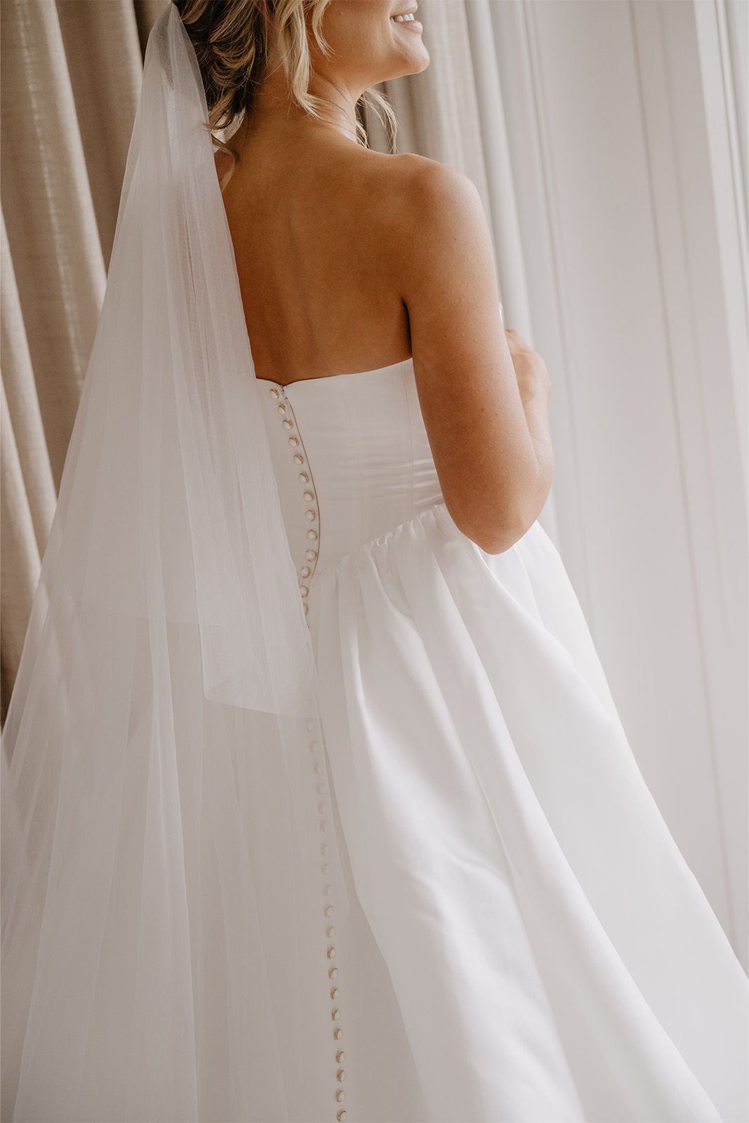 Bride in a white dress with a veil, standing by a window.