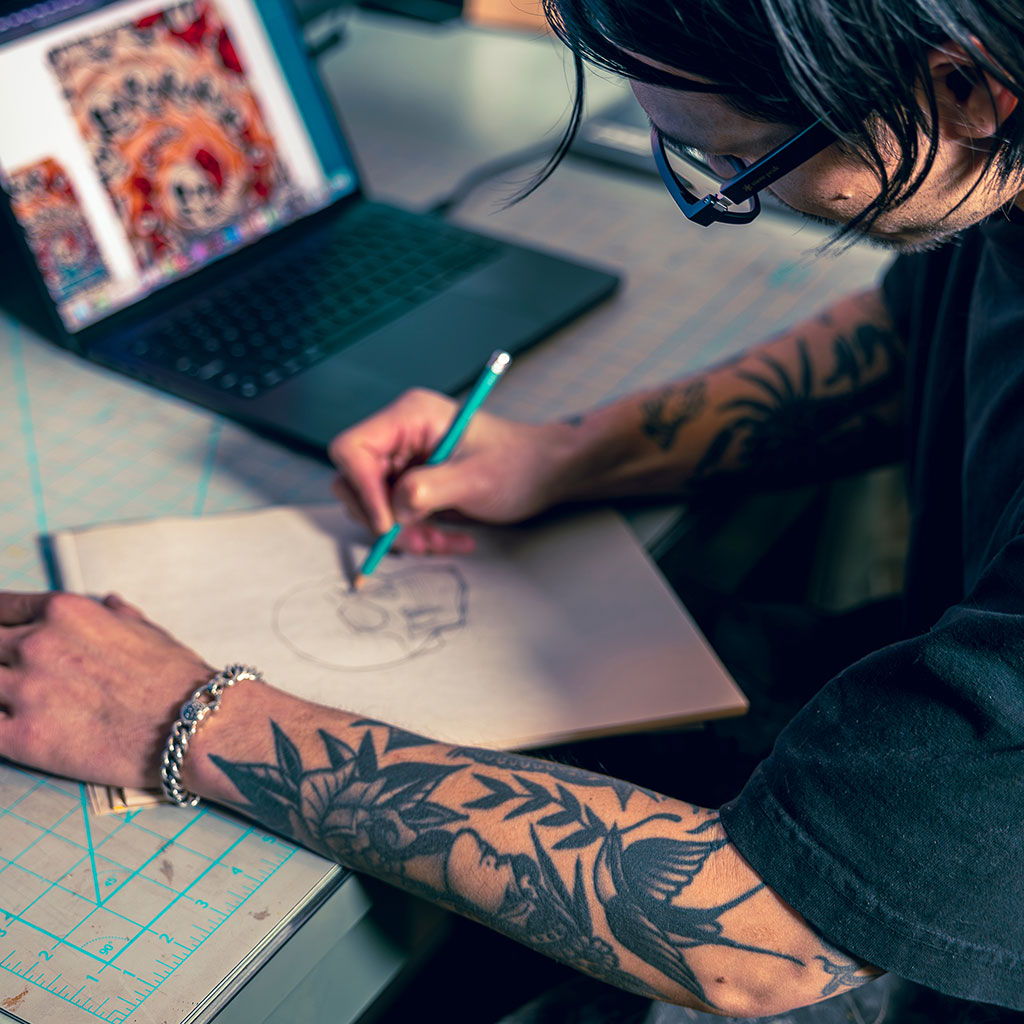 Person with tattoos sketching on paper, laptop displaying art in background.