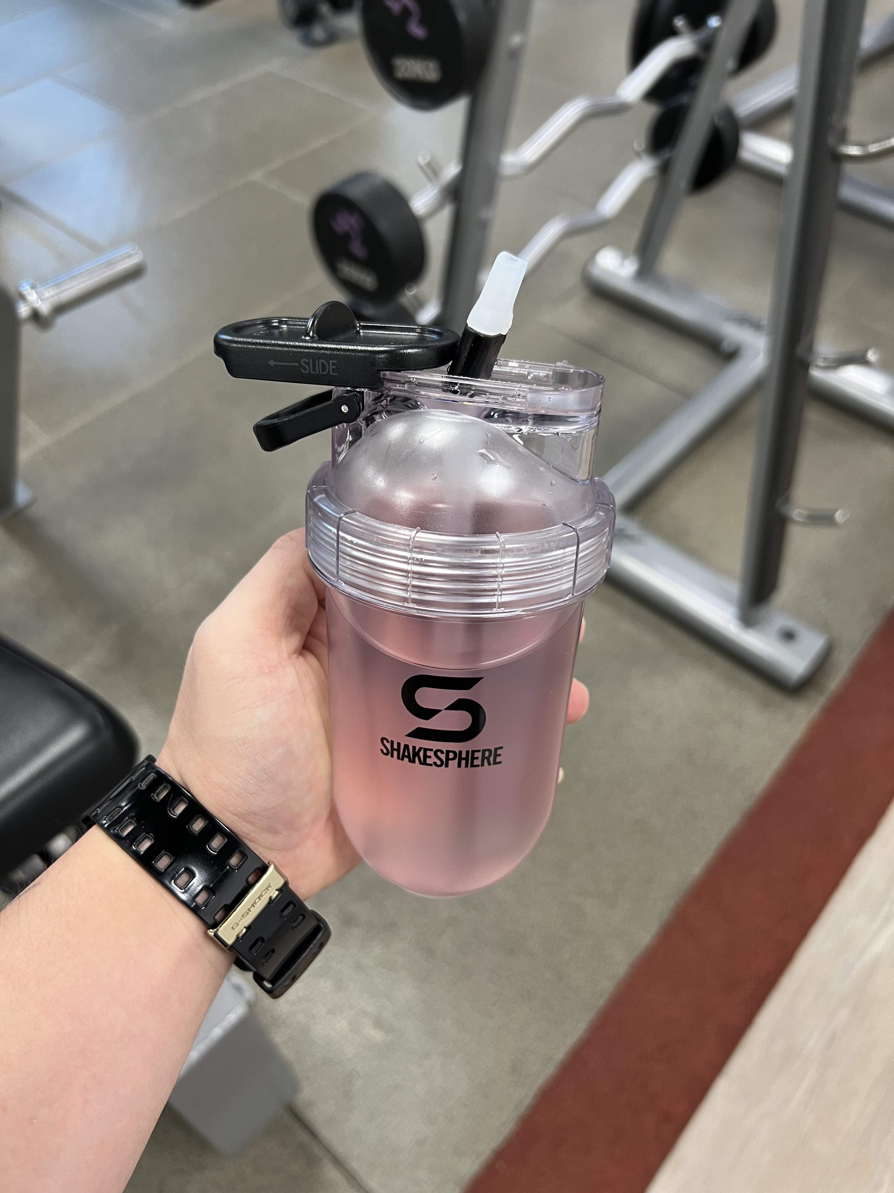 Hand holding a shaker bottle with pink liquid in a gym setting.