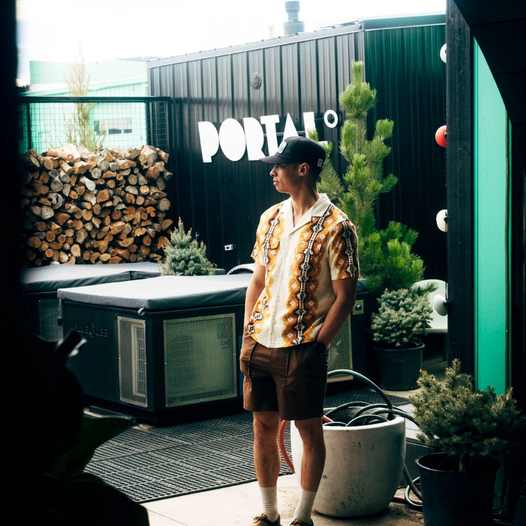 Person in a patterned shirt standing outdoors near greenery and a sign.