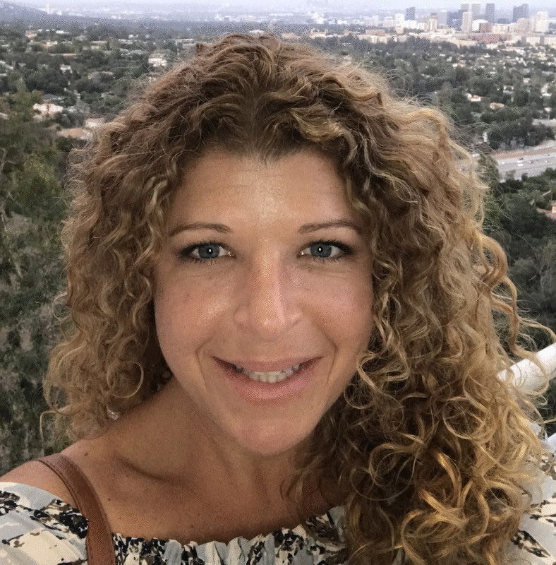 Smiling woman with curly hair against a scenic landscape in the background.