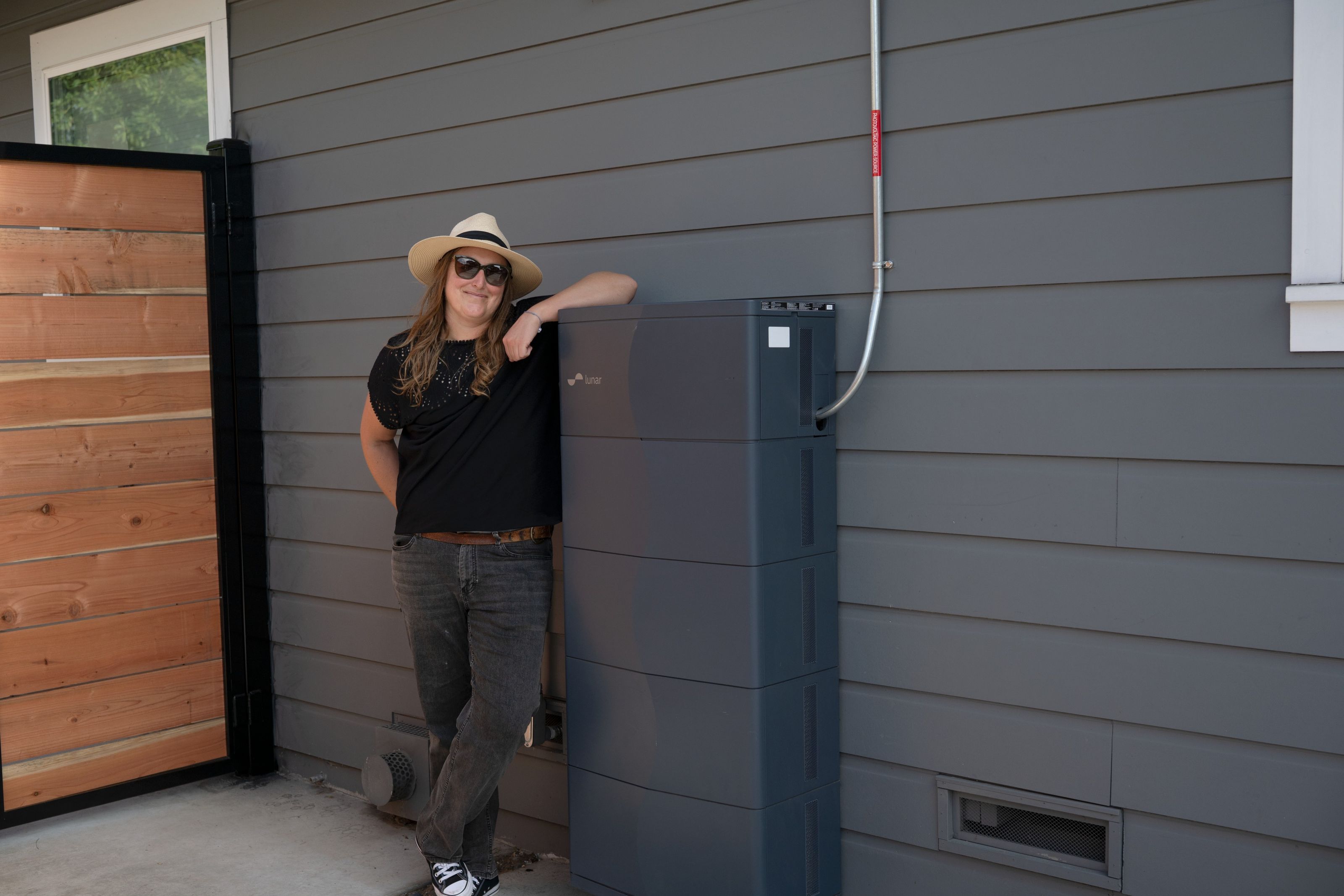 Person wearing a hat leaning against a Lunar Energy system near a wooden gate.