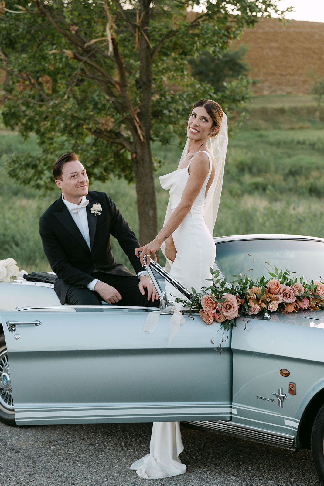 Bride and groom posed by a classic car with floral arrangements.