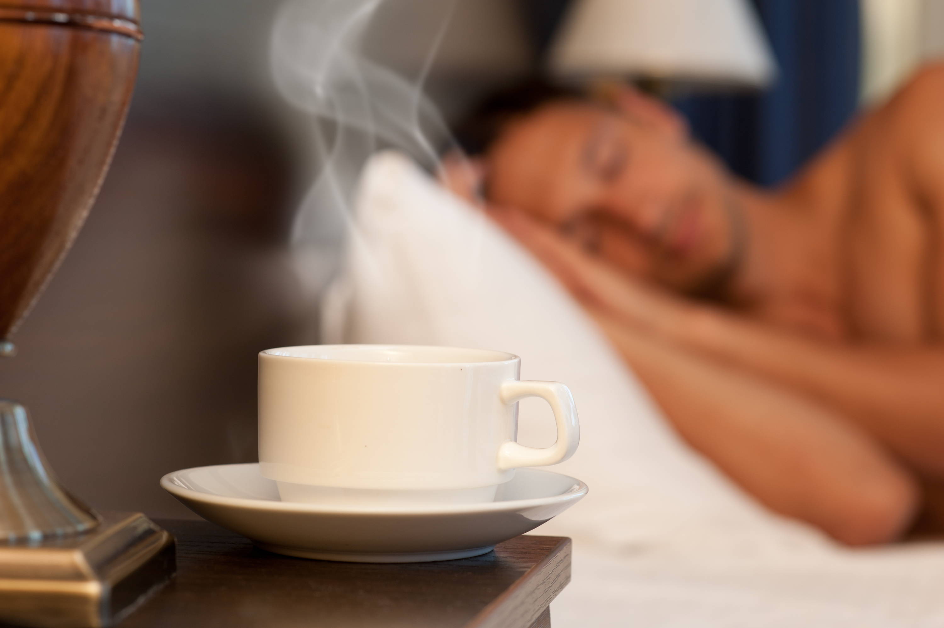A hot beverage sits on a nightstand with a man sleeping in the background.