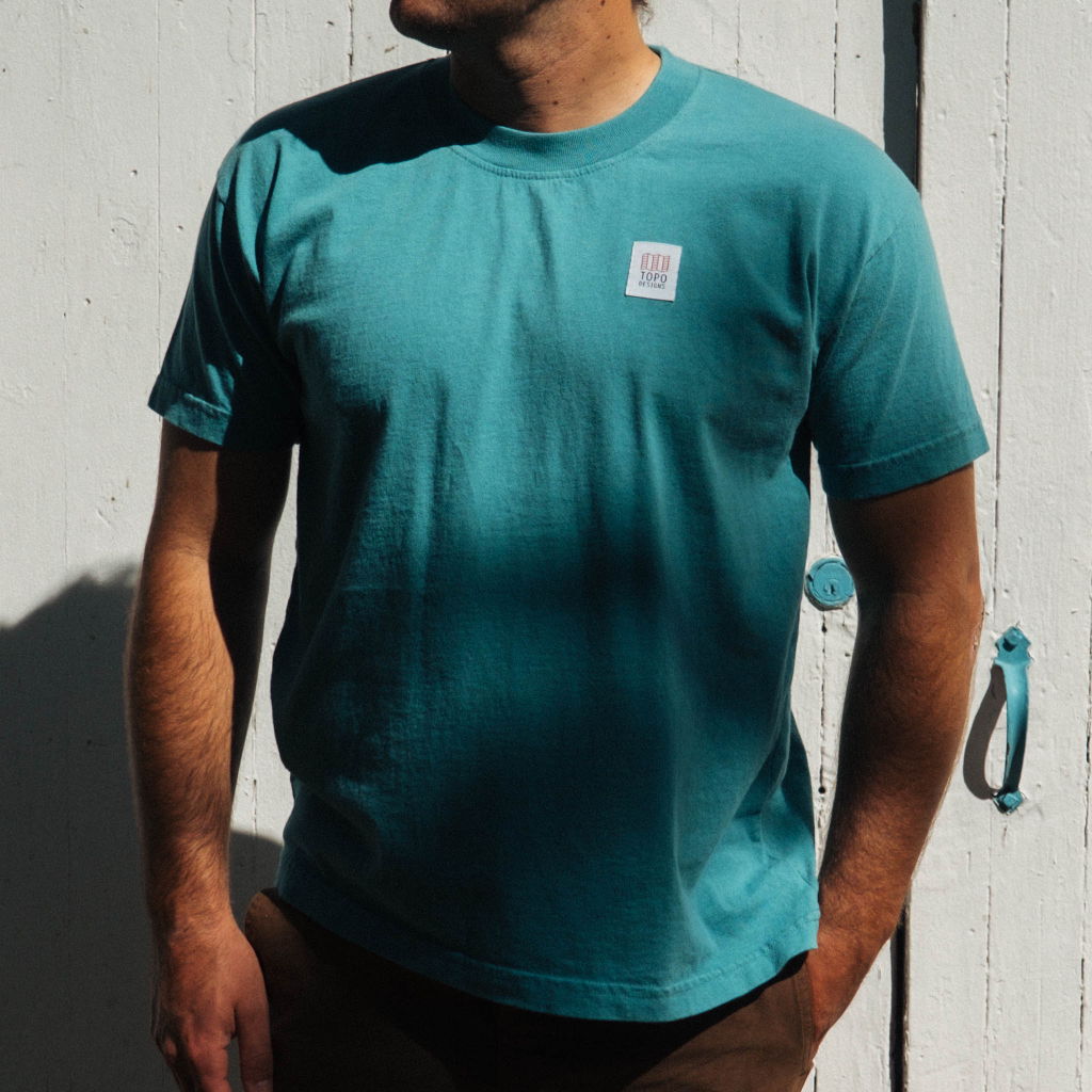 Man wearing a teal t-shirt standing against a white wall.