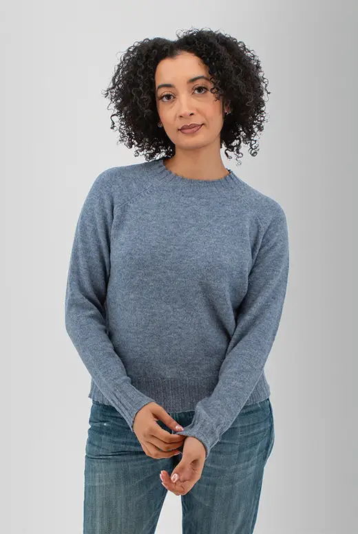 Person wearing a blue sweater and jeans with curly hair, standing against a gray background.
