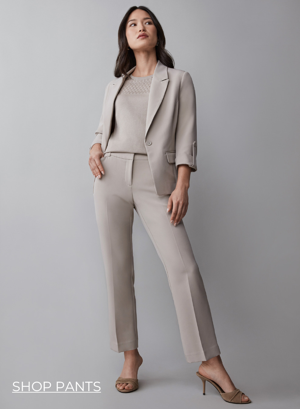 A woman in a beige pantsuit standing against a gray background.