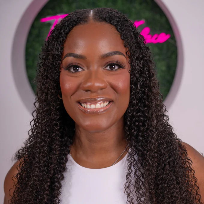 Smiling woman with curly hair extensions in front of a decorative background.
