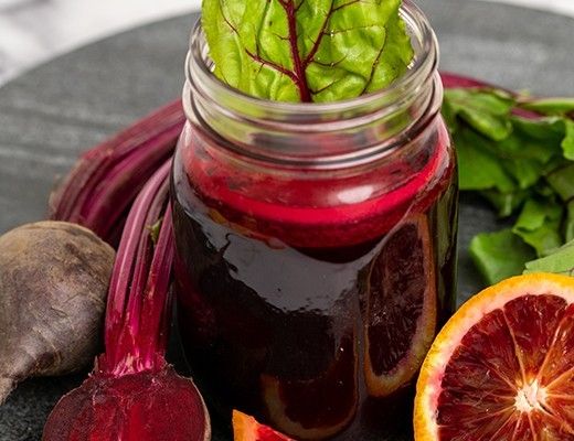 A jar of beet juice surrounded by fresh vegetables and citrus.