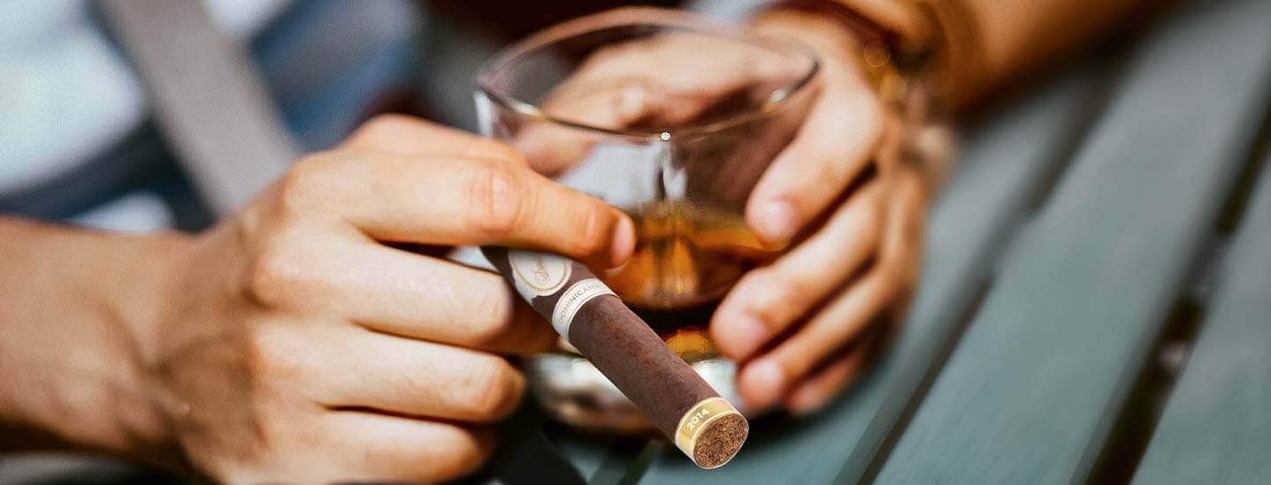 Detailaufnahme eines Mannes der eine Zigarre in der rechten Hand hält und in der linken Hand ein Glas Rum