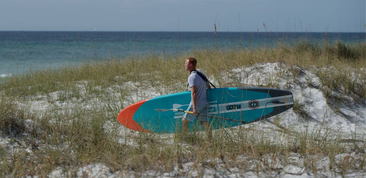 Man carrying his Flood 10′6″ Full Trax Aqua Paddle Board using the Travelink Sling