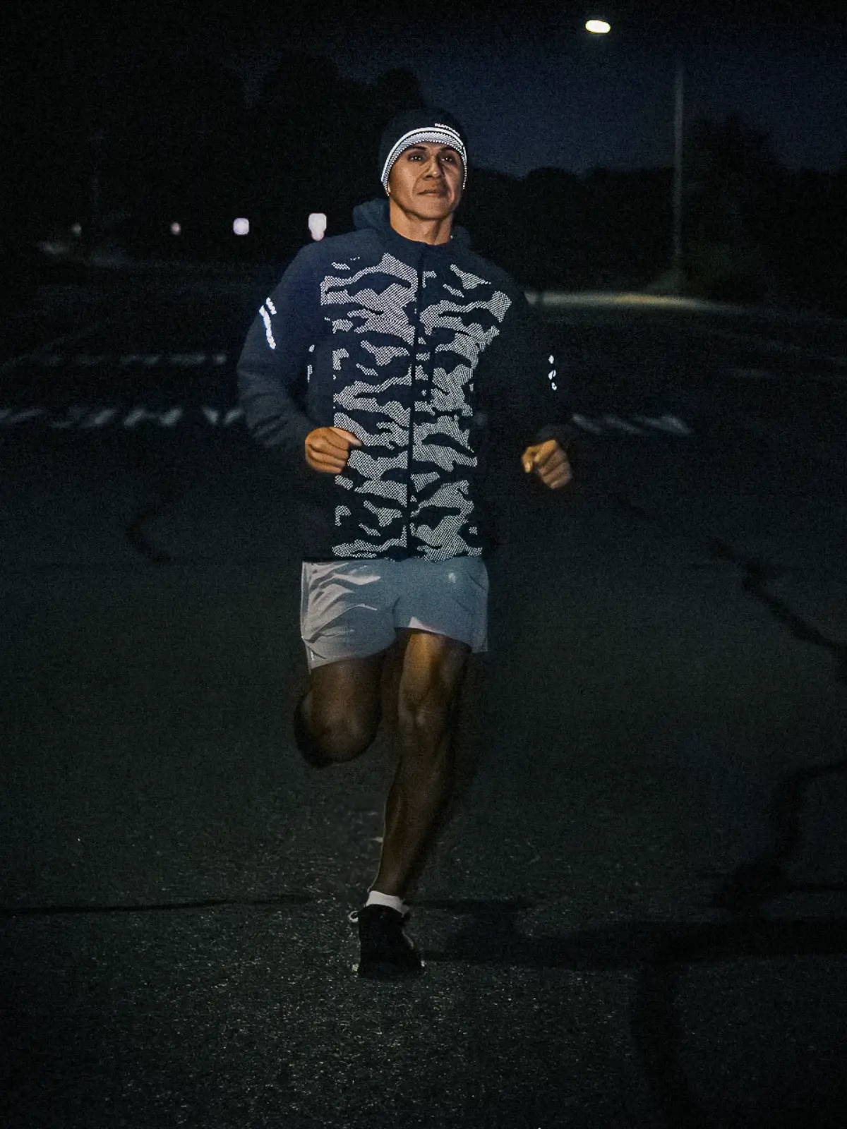 Male runner running down the street at night wearing a Nathan HyperNight Stealth Jacket which shows off the reflectivity of the jacket