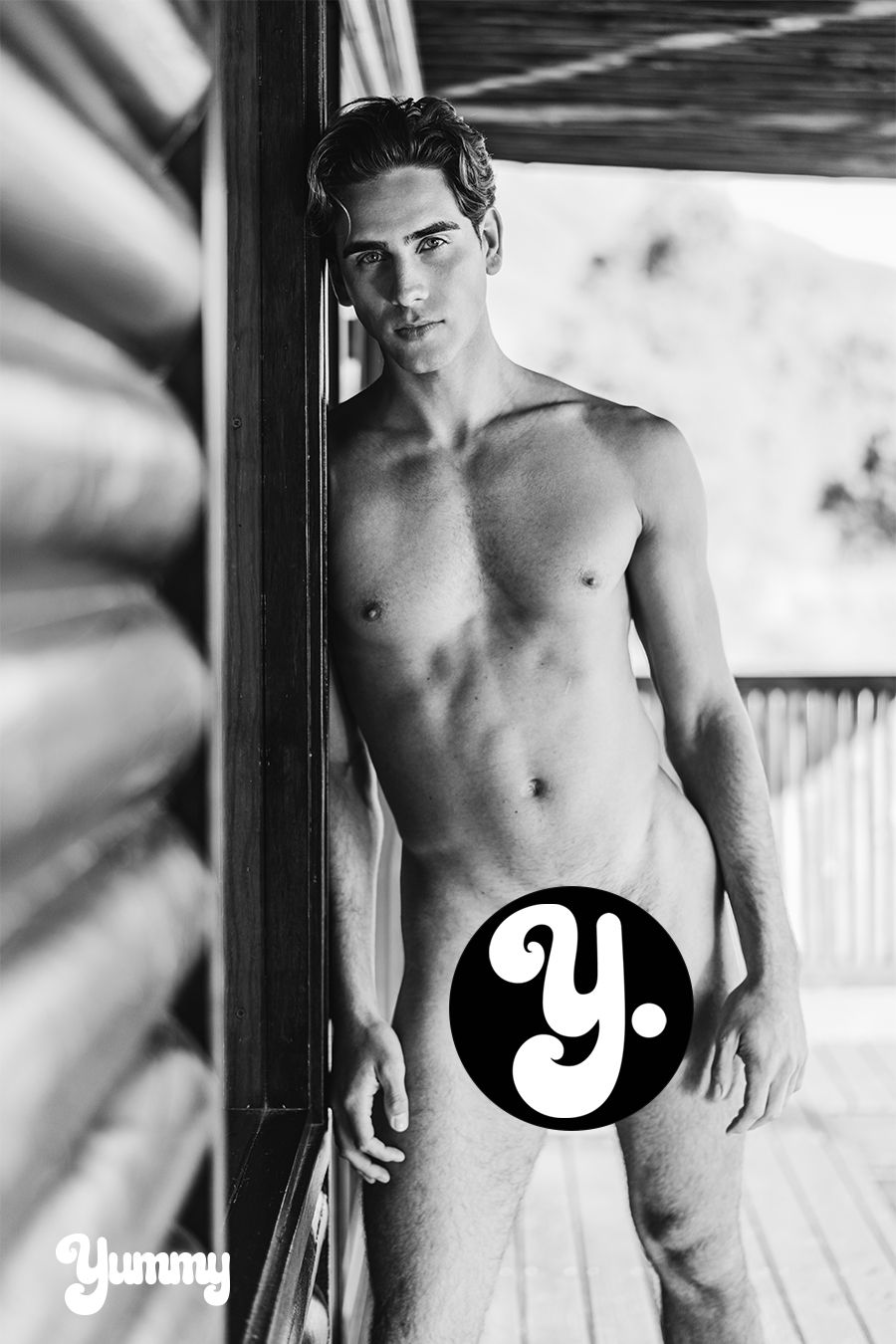 A young man posing nude against a wooden wall, partially obscured by a logo.