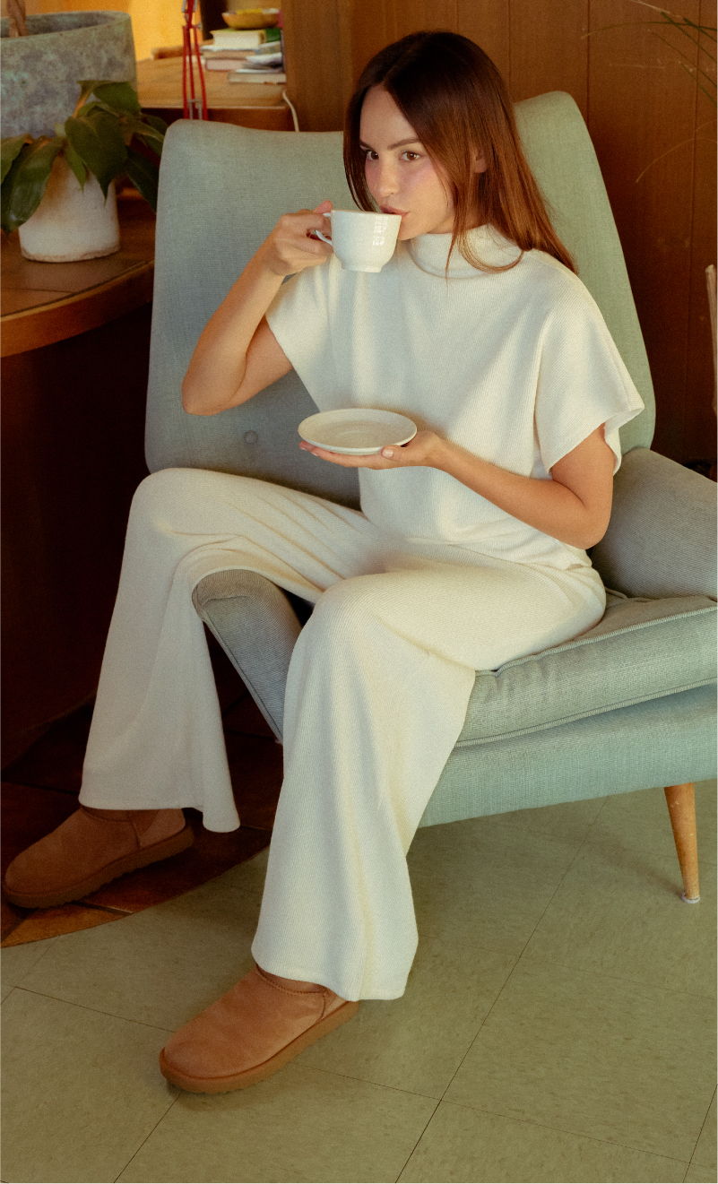 Person sitting in a chair, holding a cup and saucer, wearing white clothing.