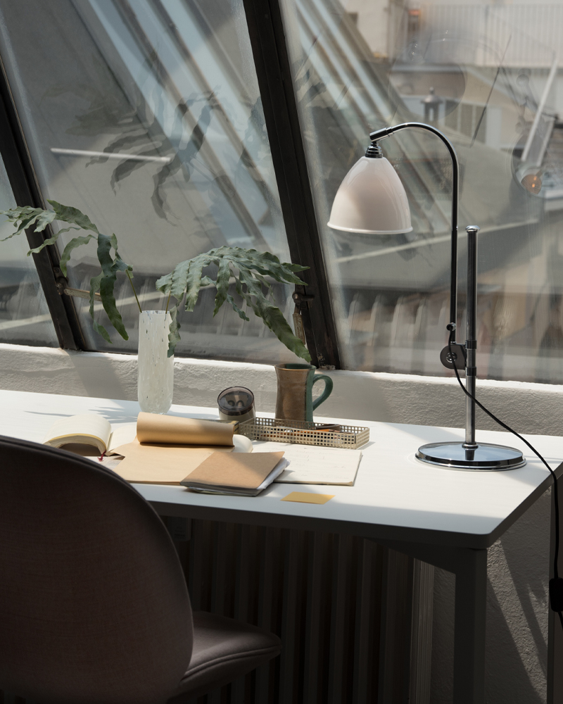 A modern Gubi desk with a BL1 table lamp, plant, and stationery by a large window.