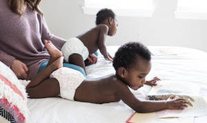 A picture of a mom with two babies, crawling on a bed with white sheets and they children are both wearing white cloth diapers. 