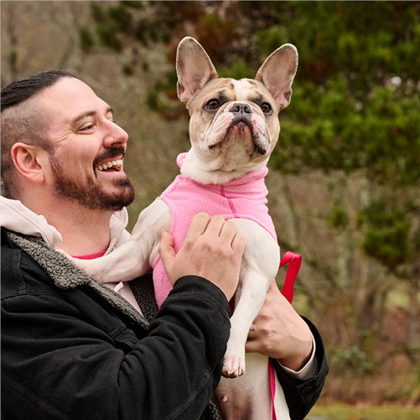 Man smiling while holding a dog in a pink outfit outdoors.