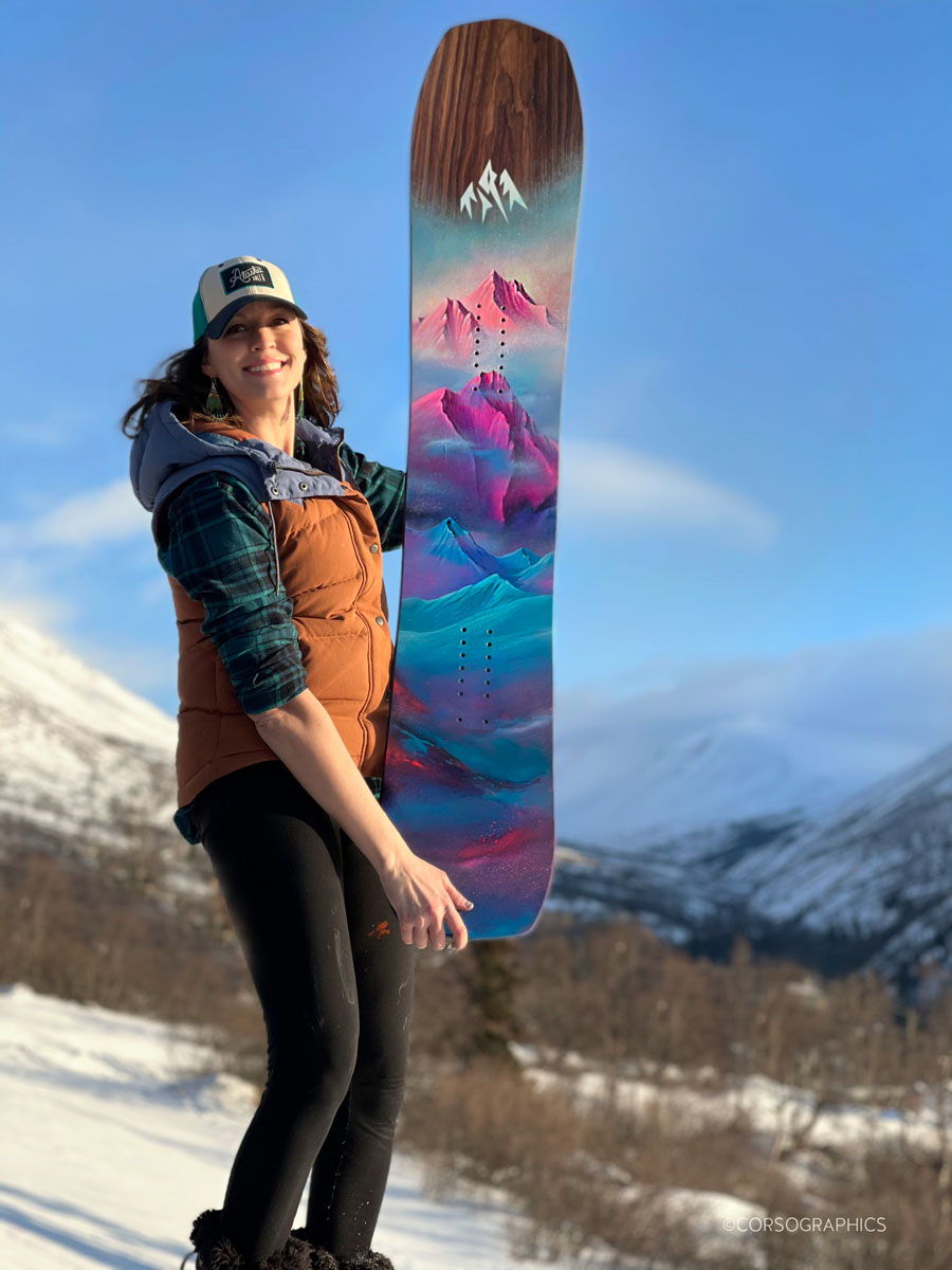 Person holding a colorful snowboard against a mountainous winter backdrop.
