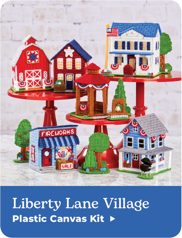 Colorful toy buildings displayed on a tiered stand with a blue background. Liberty Lane Village Plastic Canvas Kit.