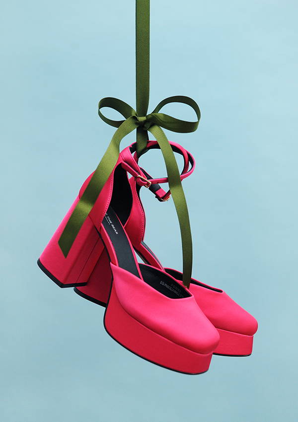 A pair of pink high heels hanging from a green ribbon bow against a blue background..