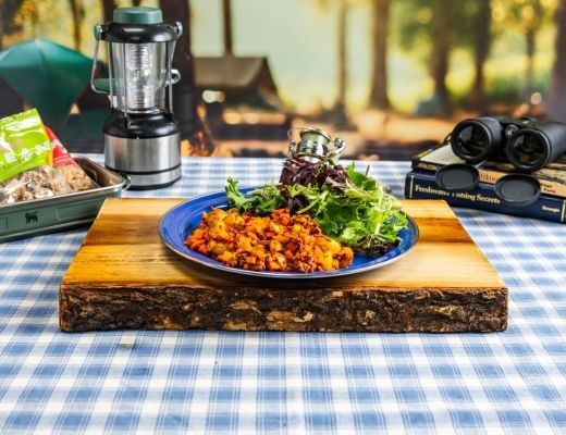Plate of food on a wooden board, with camping gear in the background.